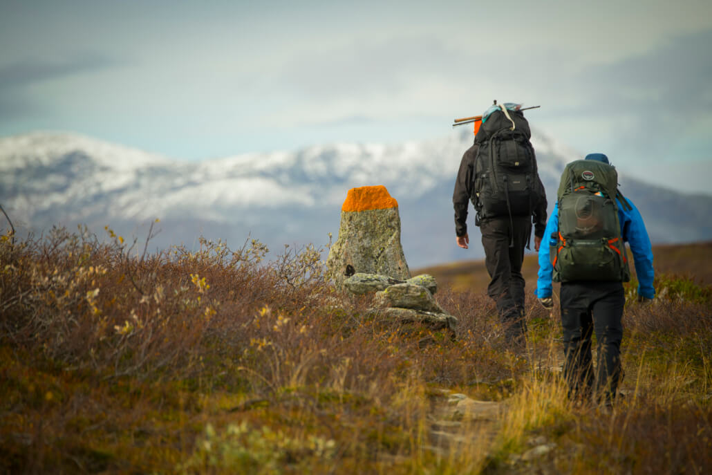 Två vandrare vandrar mot en målad sten. I bakgrunden snötäckta fjäll.