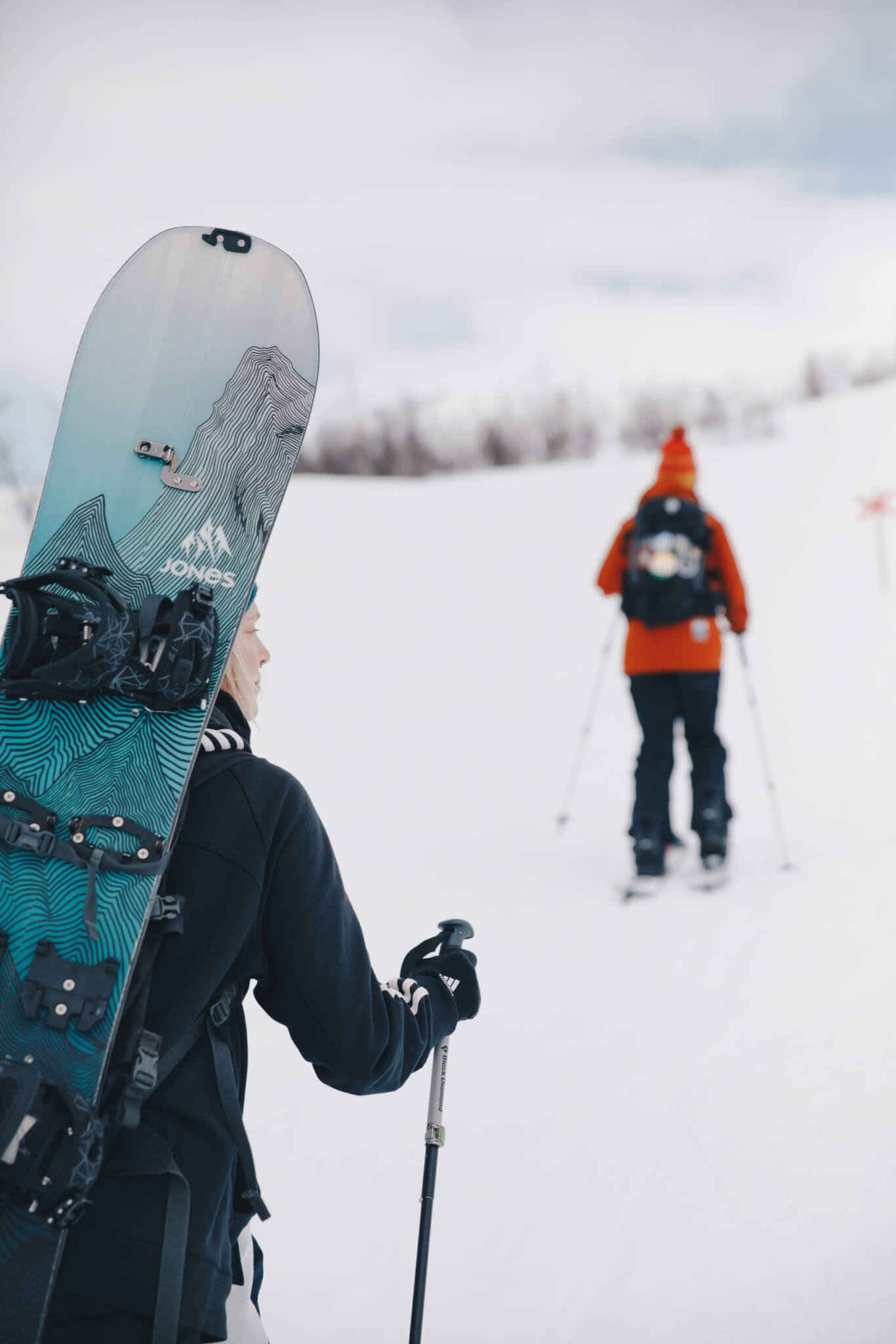 Person med snowboard på ryggen går upp för snöig backe.