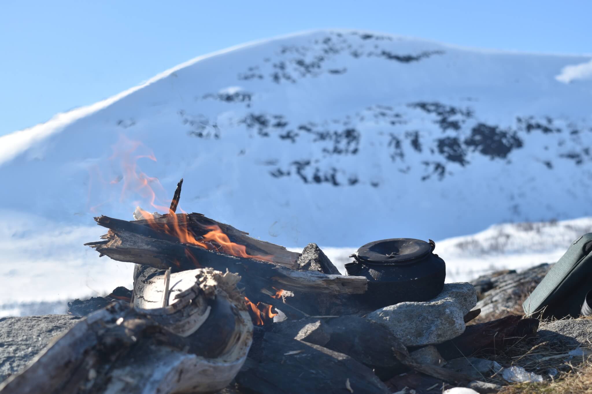 En öppen eld på kalfjäll med sotig kaffepanna bredvid. I bakgrunden stort snötäckt fjäll.