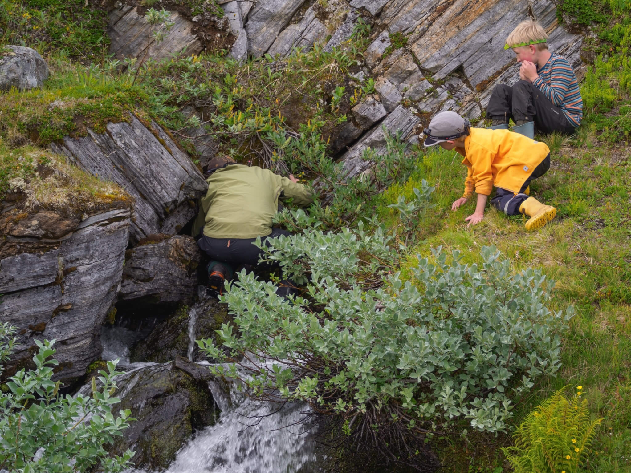 En vuxen person med grön tröja står med ryggen vänd mot kameran och tittar in i en bergskreva. Ett barn står på sidan och tittar på den vuxne.