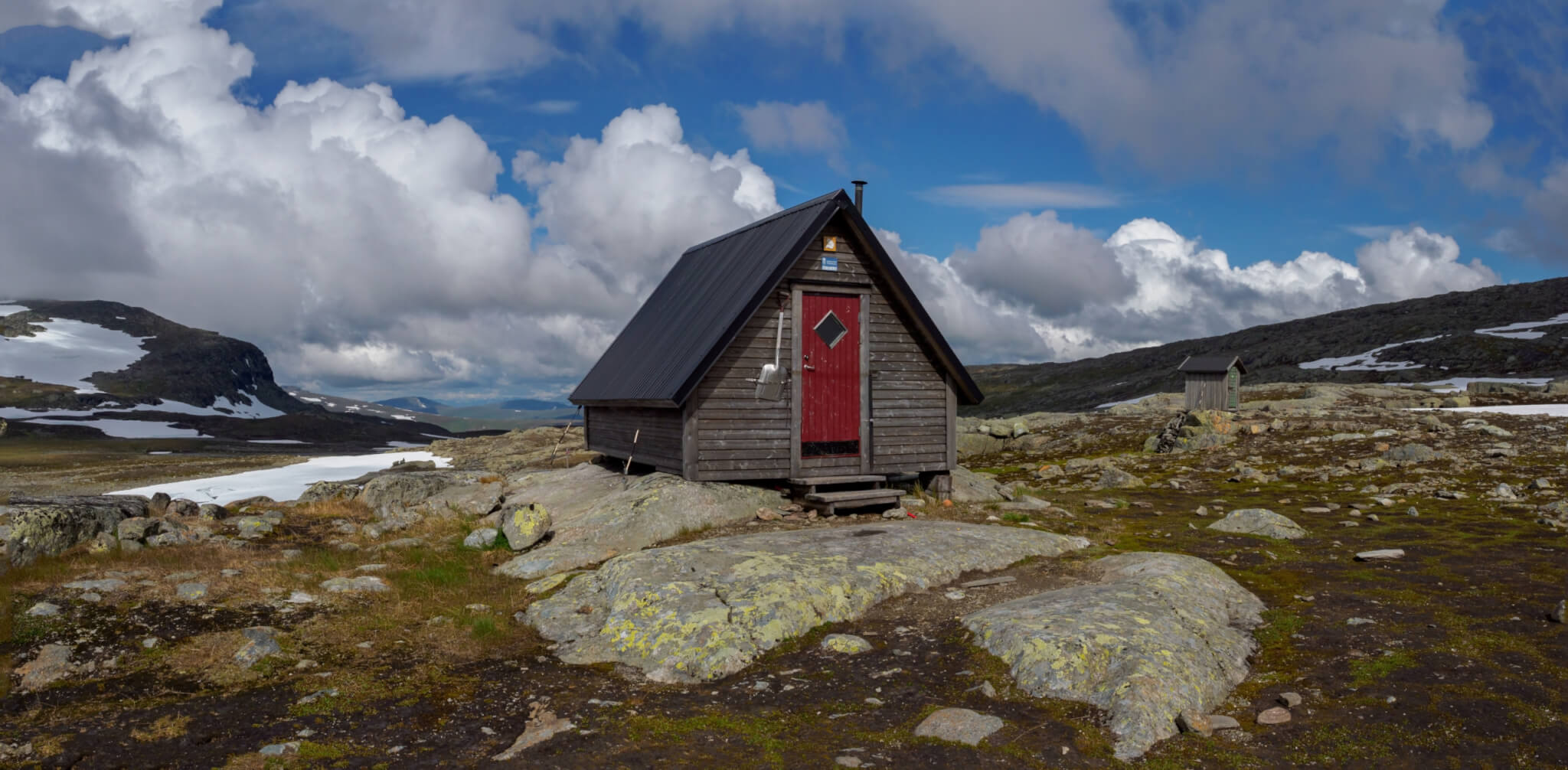 Vallentjåkke/Marinstugan. En grå liten stuga med röd dörr uppe på kalfjäll med blå himmel och moln i bakgrunden. 