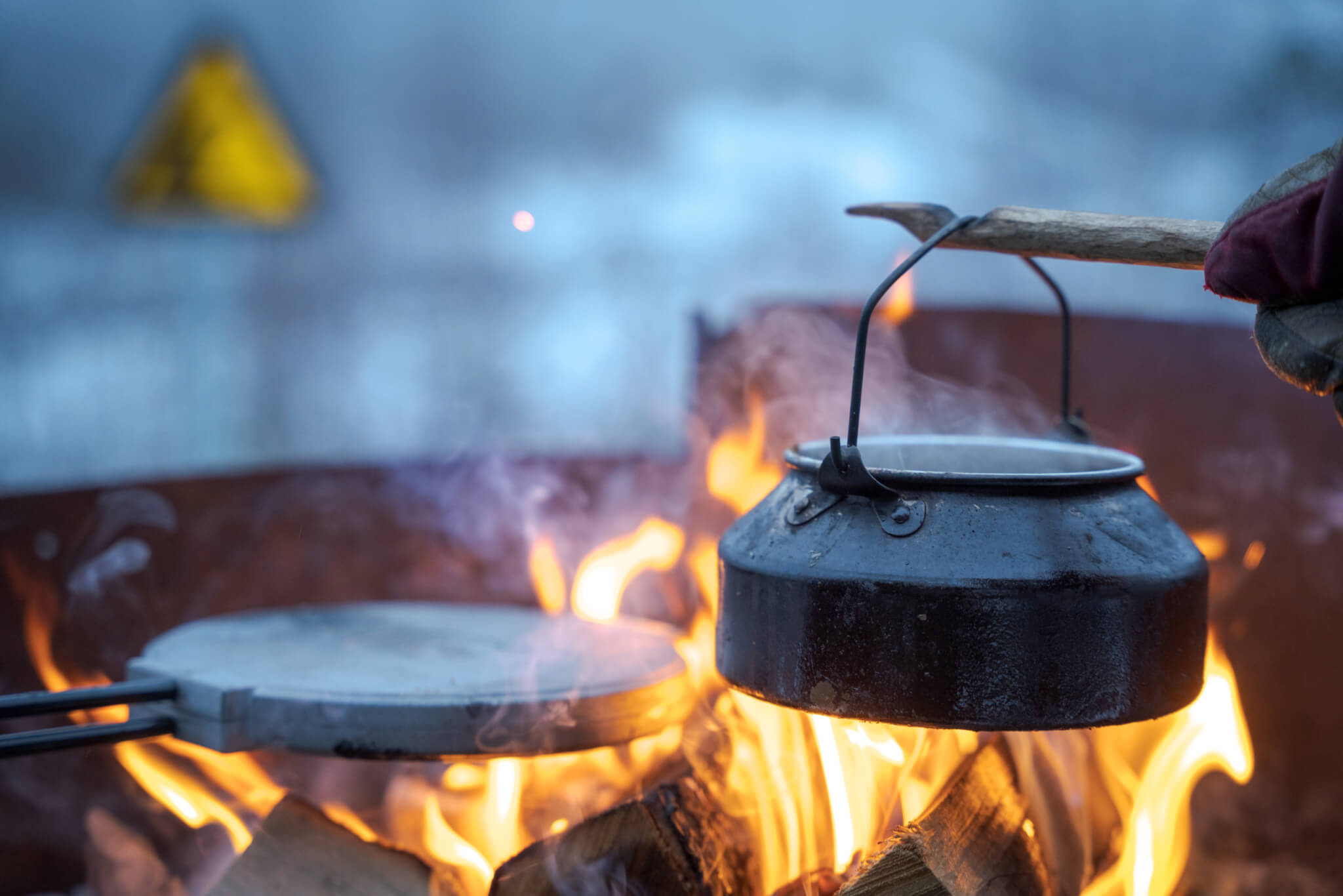 En kaffepanna över öppen eld. Tillvänster i elden syns ett våffeljärn. I bakgrunden syns en gul triangelskylt suddigt.