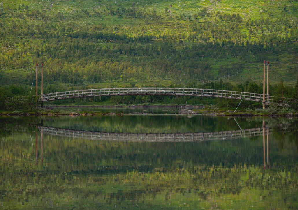 Tärnasjön. Bro över spegelblank sjö. Grön lövskog speglas i sjön.