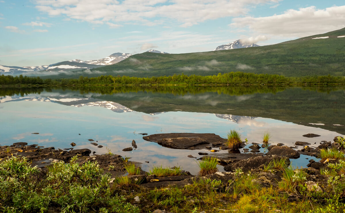 Tärnasjön ligger spegelblank en solig sommardag. Runda fjäll speglar sig i vattnet.