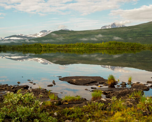 Tärnasjön ligger spegelblank en solig sommardag. Runda fjäll speglar sig i vattnet.